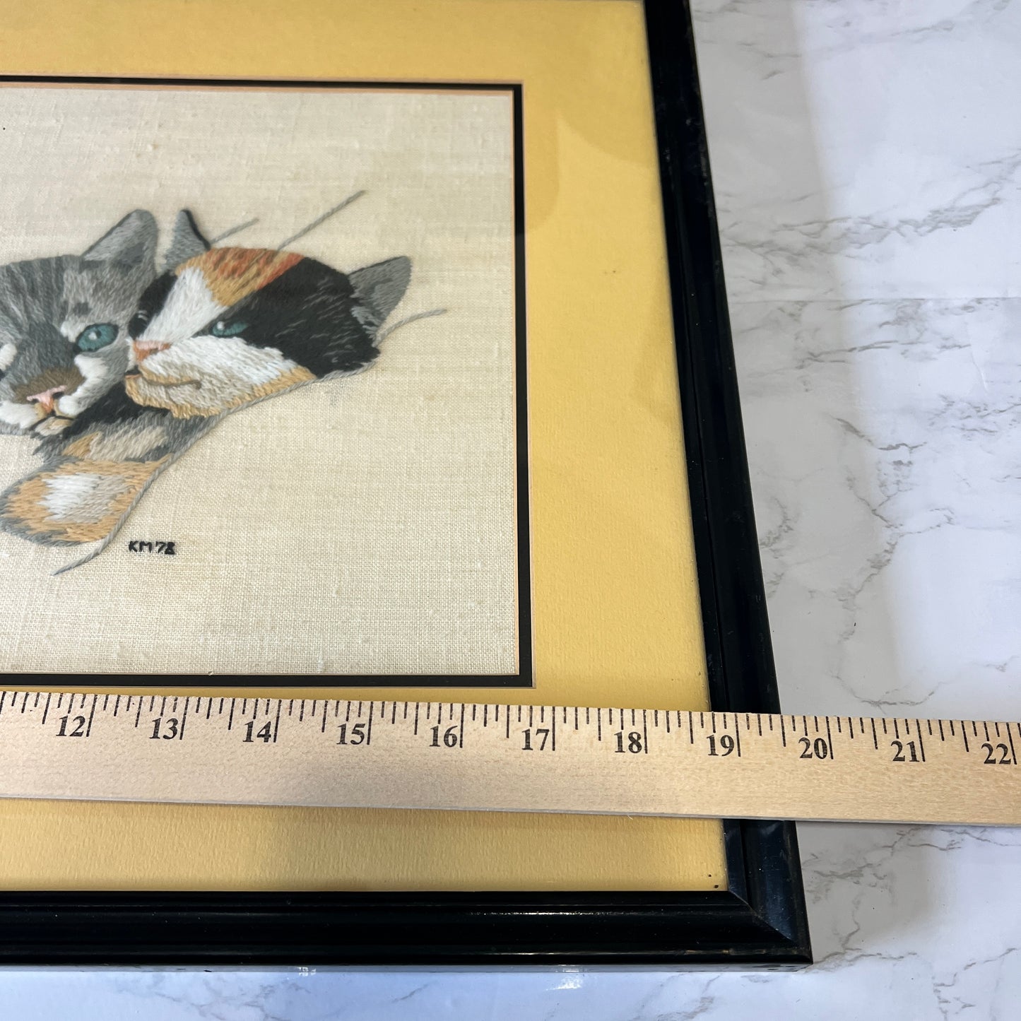 A framed embroidered cat artwork with a black frame and pale yellow mat, shown on a marble-like surface with a wooden ruler across the bottom to indicate size.
