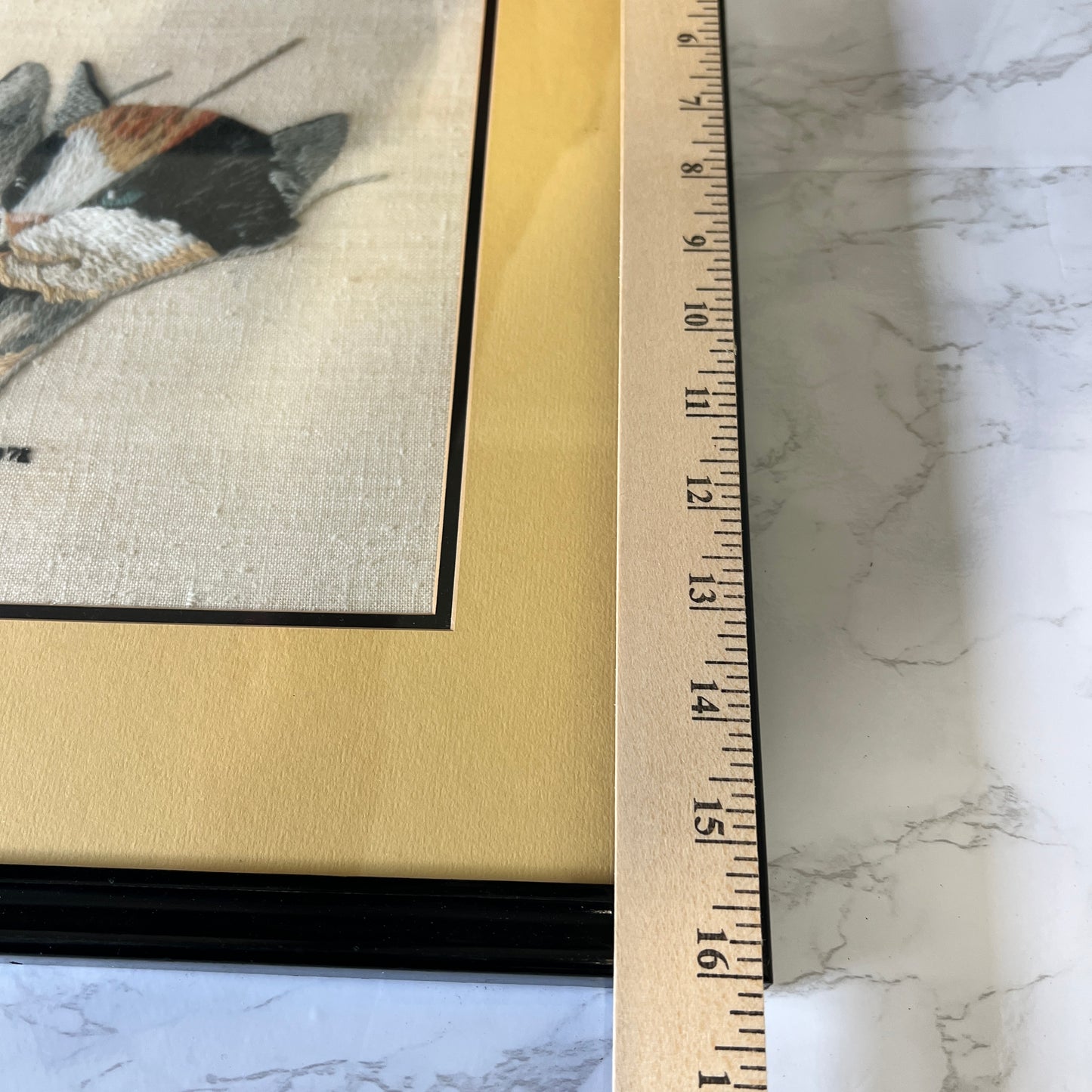 Close-up of a framed art print showing a butterfly or moth illustration on textured off-white fabric, with a gold mat and a wooden ruler standing vertically to indicate scale, placed on a white marble surface.