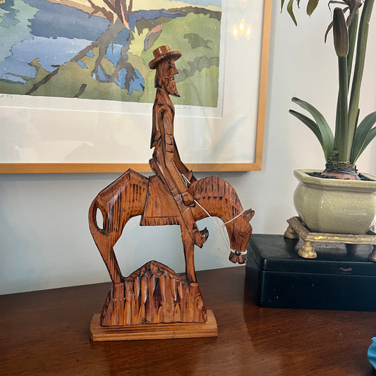 Wooden sculpture of a cowboy on horseback on a wooden surface with a painting and plant in the background.