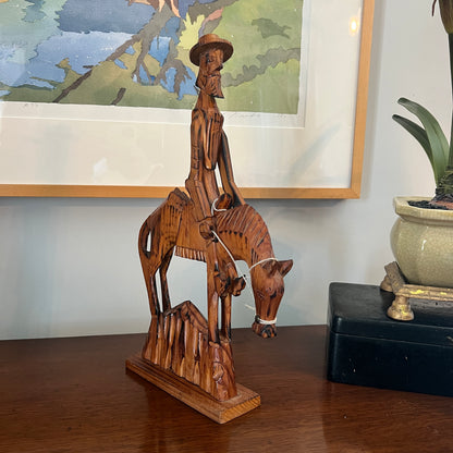 Wooden sculpture of a person on horseback on a wooden surface with a framed picture in the background.