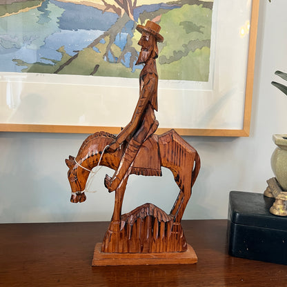 Wooden sculpture of a cowboy on horseback with a framed painting in the background.