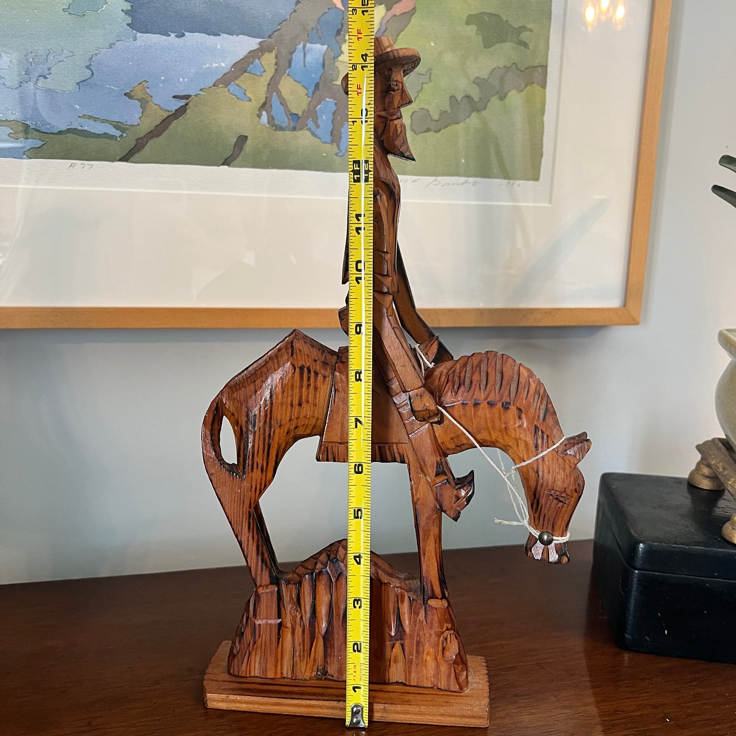 Wooden sculpture of a man on horse with a tape measure for scale, against a wall with framed artwork.