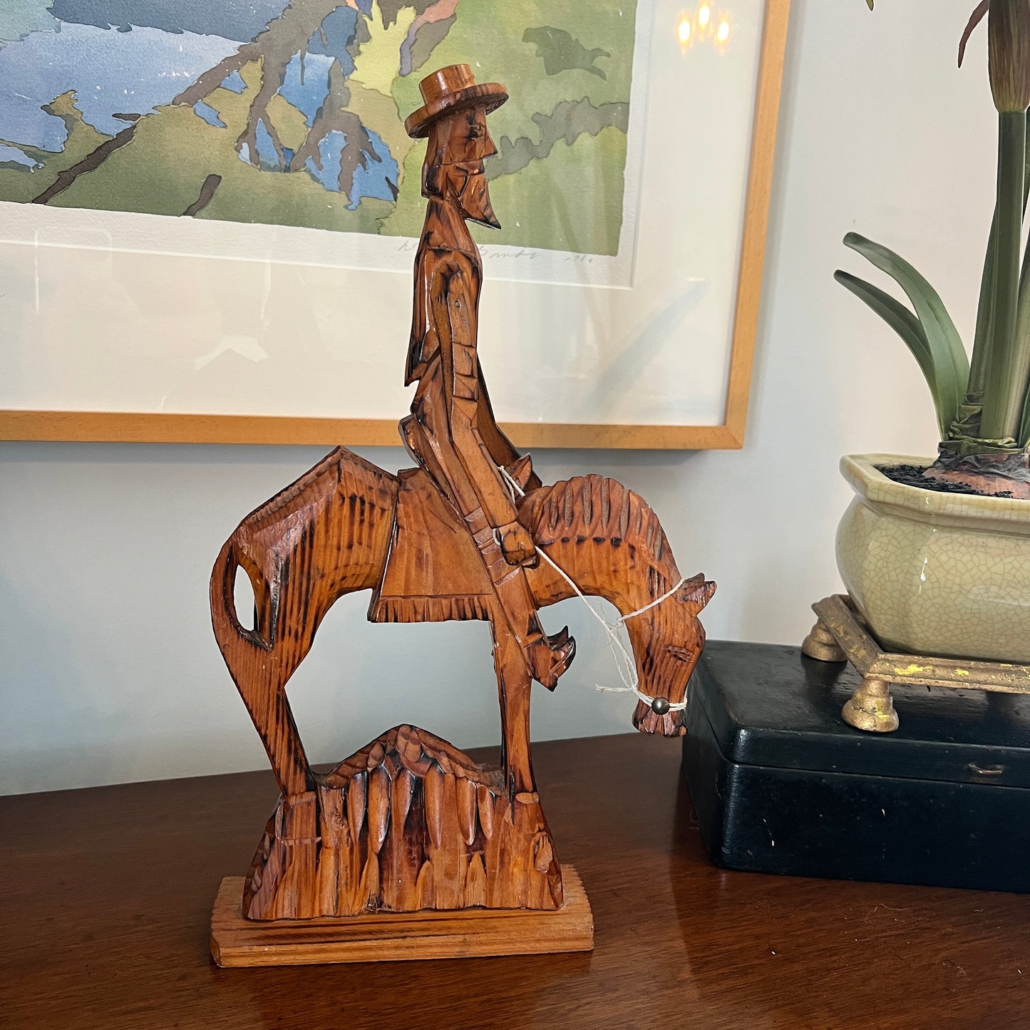 Wooden sculpture of a cowboy on horseback on a table with a painting and plant in the background.