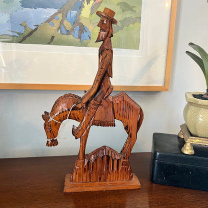 Wooden sculpture of a man riding a horse on a table with a framed picture and plant in the background.