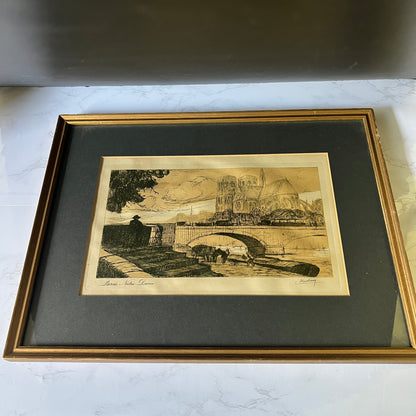 A framed art print depicting a historic stone bridge over water with buildings in the background, mounted with a dark gray mat and a gold-toned frame on a light marble surface.