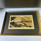 A framed art print depicting a historic stone bridge over water with buildings in the background, mounted with a dark gray mat and a gold-toned frame on a light marble surface.
