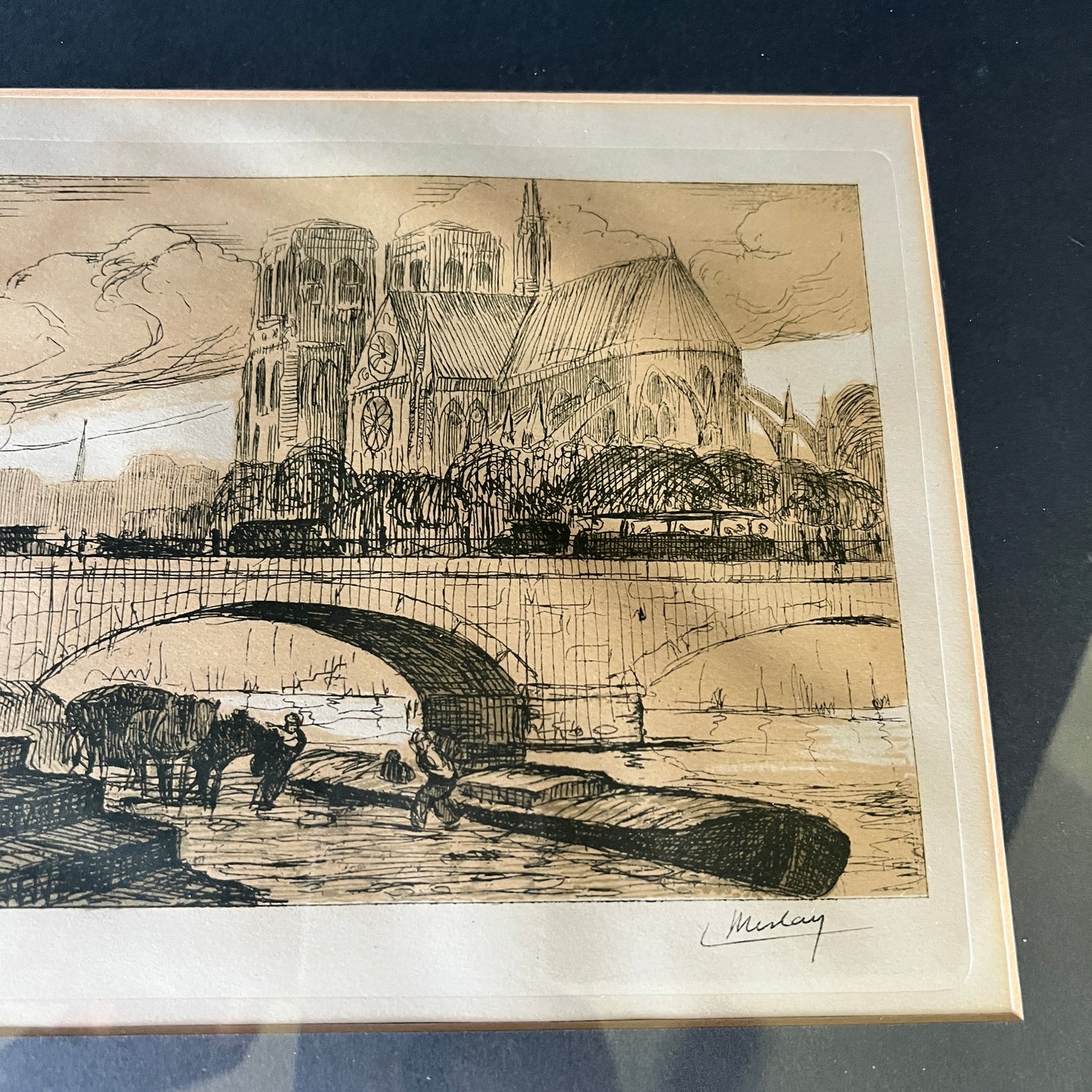 A framed sepia-toned art print depicting a cathedral and bridge scene, photographed from above on a dark surface. The print has a cream border and a visible artist signature in the lower right.