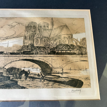A framed sepia-toned art print depicting a cathedral and bridge scene, photographed from above on a dark surface. The print has a cream border and a visible artist signature in the lower right.