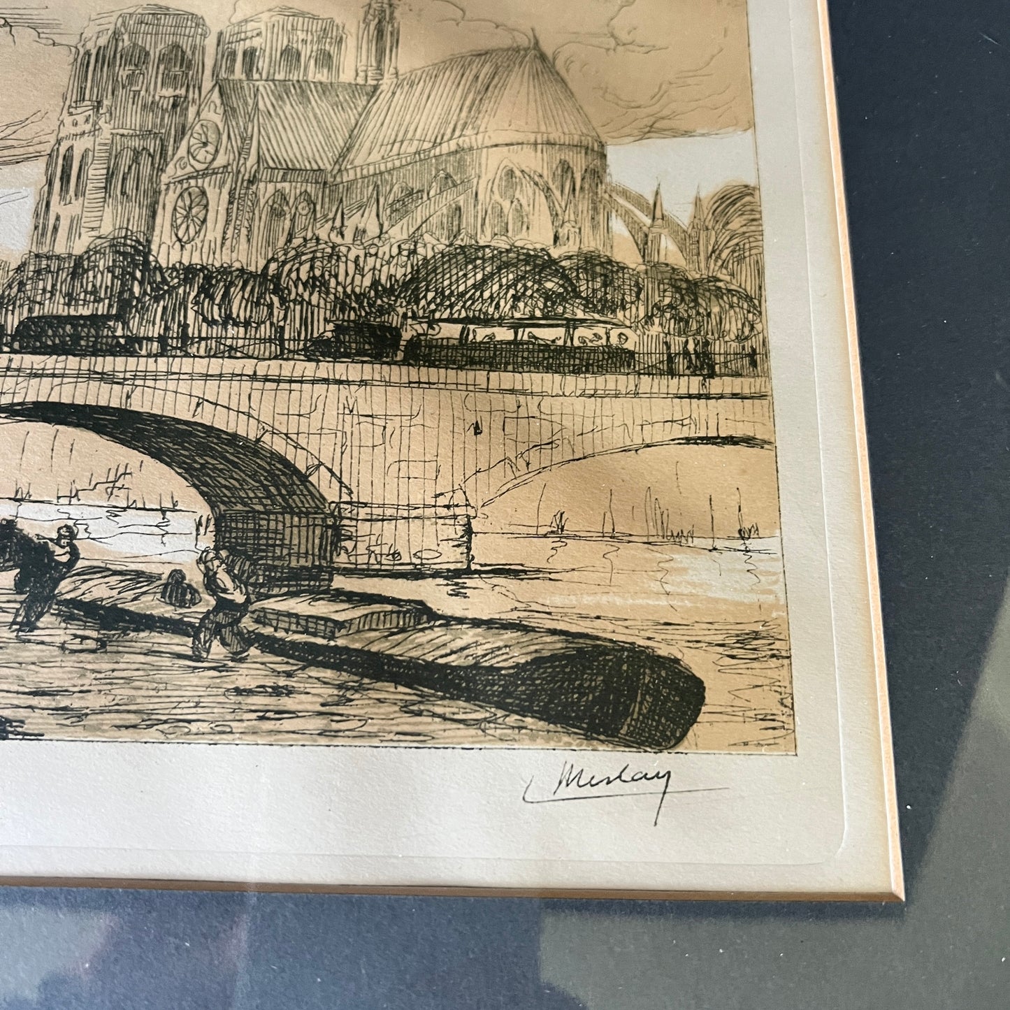 A framed monochrome art print depicting a Parisian scene with Notre Dame Cathedral, a bridge, and a boat in the foreground, signed by the artist. The print is mounted on a light beige mat and shown resting on a dark reflective surface.