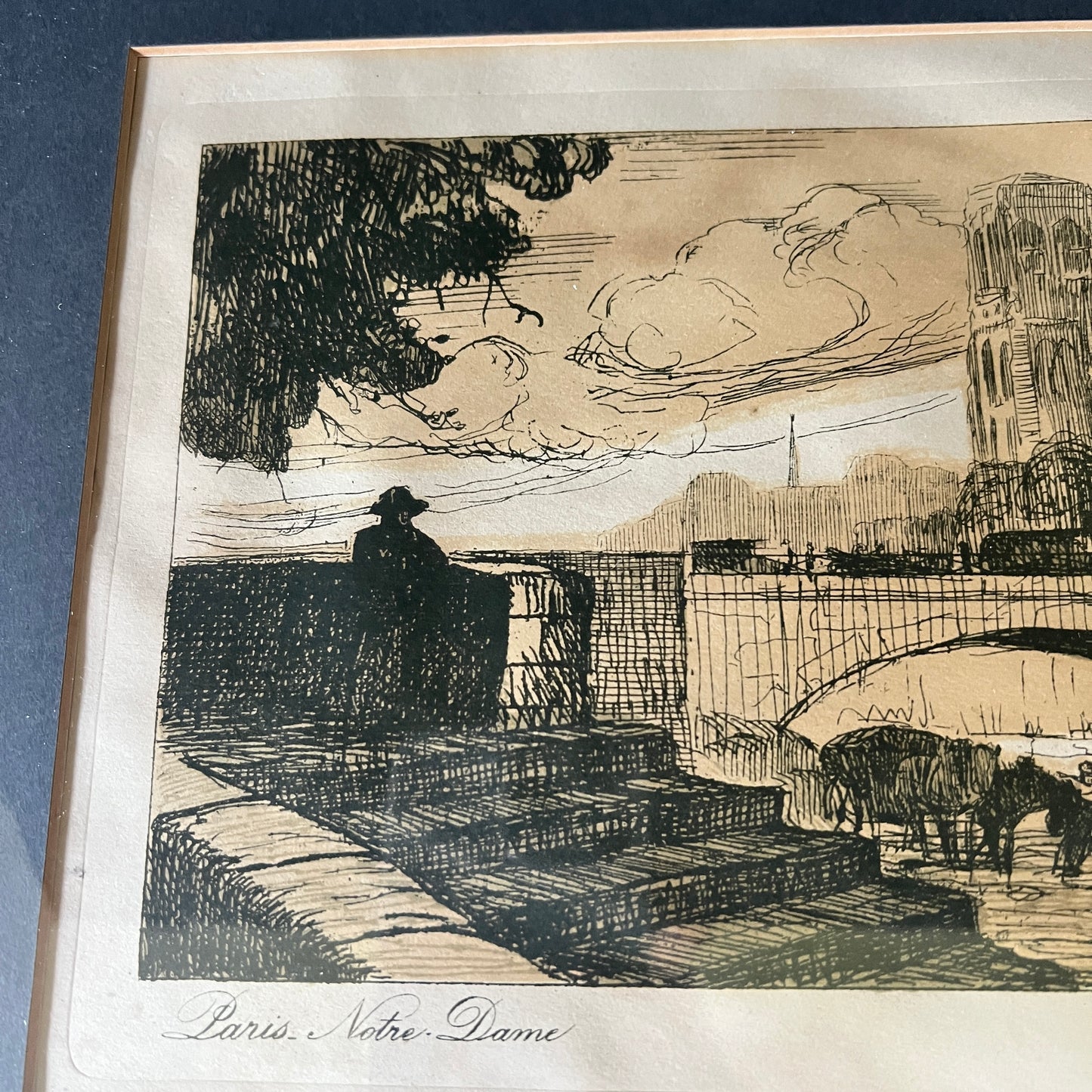 A framed black ink sketch print on aged beige paper, depicting a city scene with a bridge, a silhouetted figure, trees, and a tower. The print is mounted with a wide white mat and a light wood frame, photographed against a dark blue background.