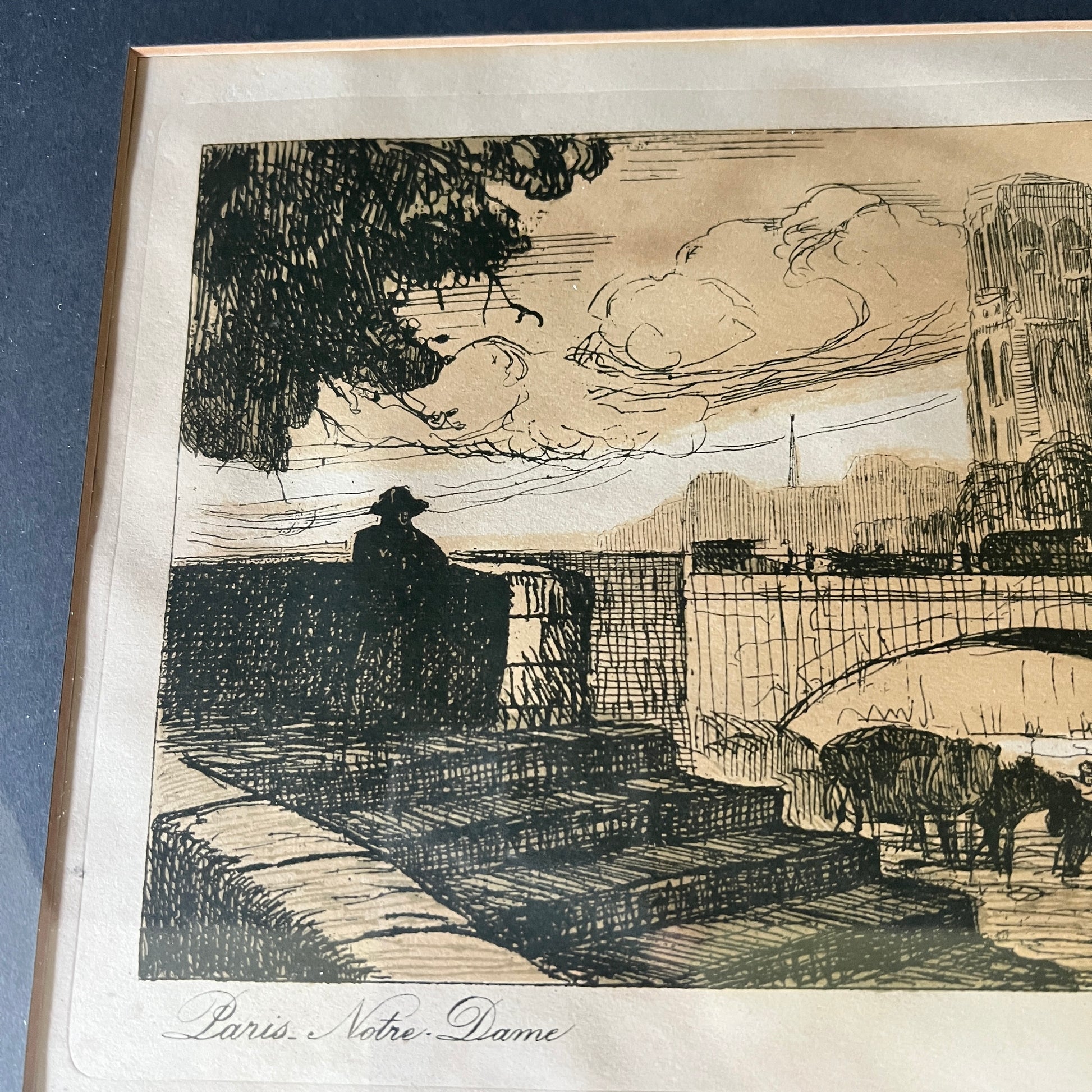 A framed black ink sketch print on aged beige paper, depicting a city scene with a bridge, a silhouetted figure, trees, and a tower. The print is mounted with a wide white mat and a light wood frame, photographed against a dark blue background.