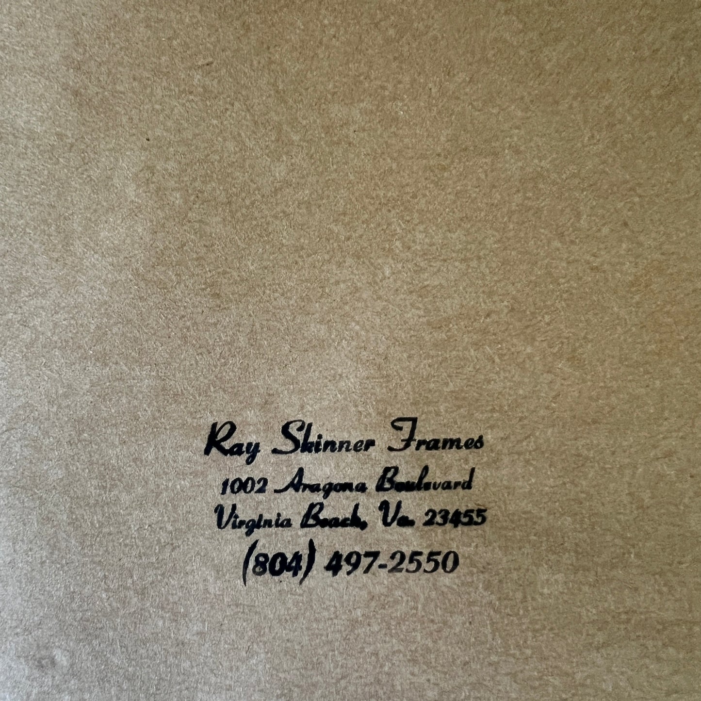 Close-up of a textured tan or kraft-paper surface with black printed text reading 'Ray Shinner Frames' and a business address/phone number in Virginia Beach.