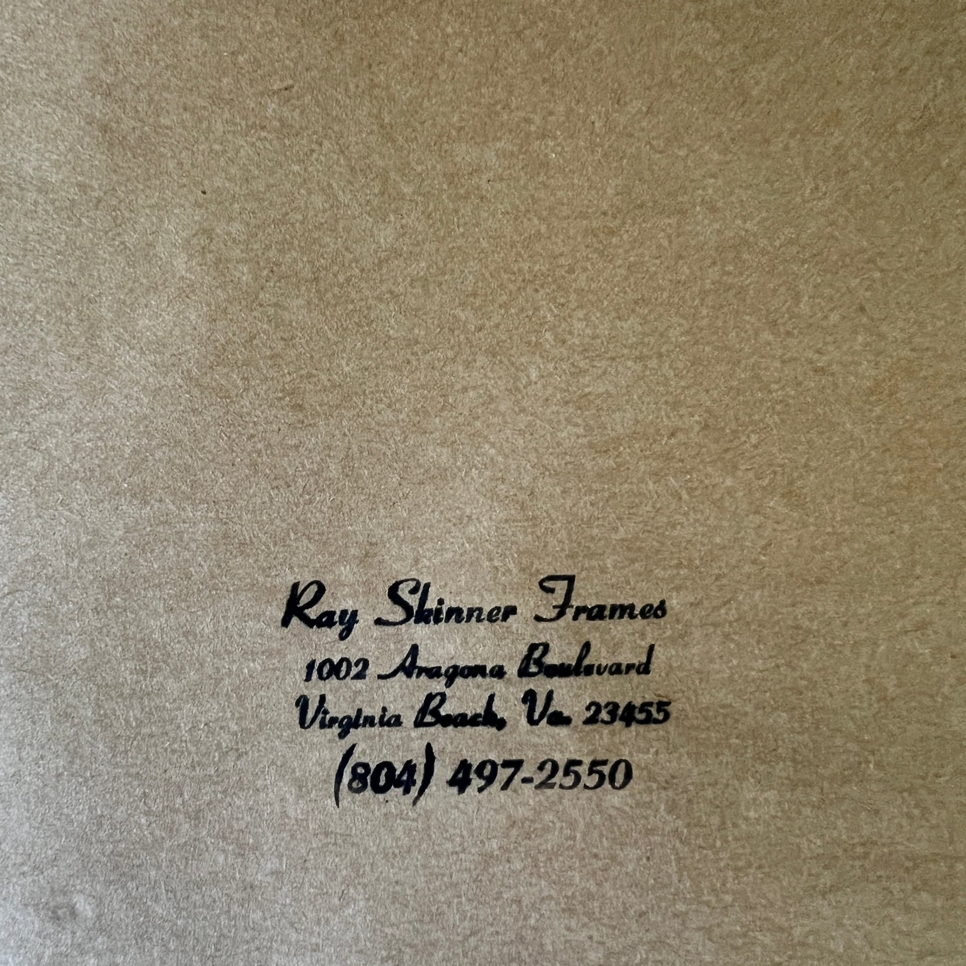 Close-up of a textured tan or kraft-paper surface with black printed text reading 'Ray Shinner Frames' and a business address/phone number in Virginia Beach.