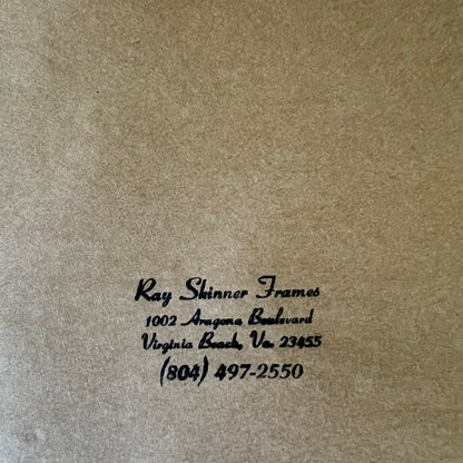 Close-up of a textured tan or kraft-paper surface with black printed text reading 'Ray Shinner Frames' and a business address/phone number in Virginia Beach.