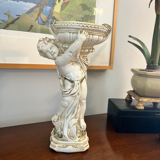 Decorative statue of a cherub holding a bowl on a wooden surface with a painting in the background.