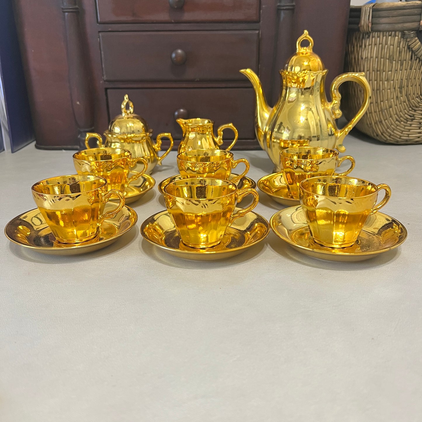 A set of shiny gold tea or coffee cups with saucers, a matching teapot, and a creamer, arranged on a light gray surface with a dark wooden cabinet in the background.