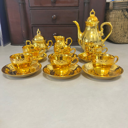A set of shiny gold tea or coffee cups with saucers, a matching teapot, and a creamer, arranged on a light gray surface with a dark wooden cabinet in the background.