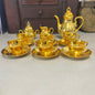 A set of shiny gold tea or coffee cups with saucers, a matching teapot, and a creamer, arranged on a light gray surface with a dark wooden cabinet in the background.