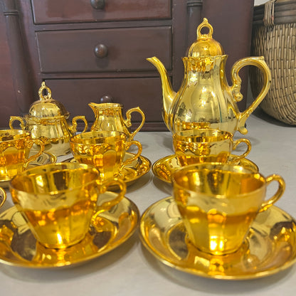 A shiny gold tea or coffee cup set with a matching teapot, creamer, and saucers, arranged on a tabletop in front of a dark wooden cabinet.