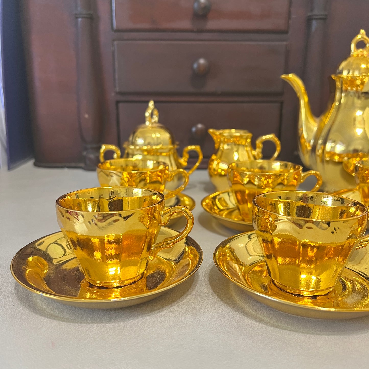 A set of shiny gold teacups with matching saucers, a gold teapot, and a gold creamer, arranged on a tabletop with a dark wooden cabinet in the background.