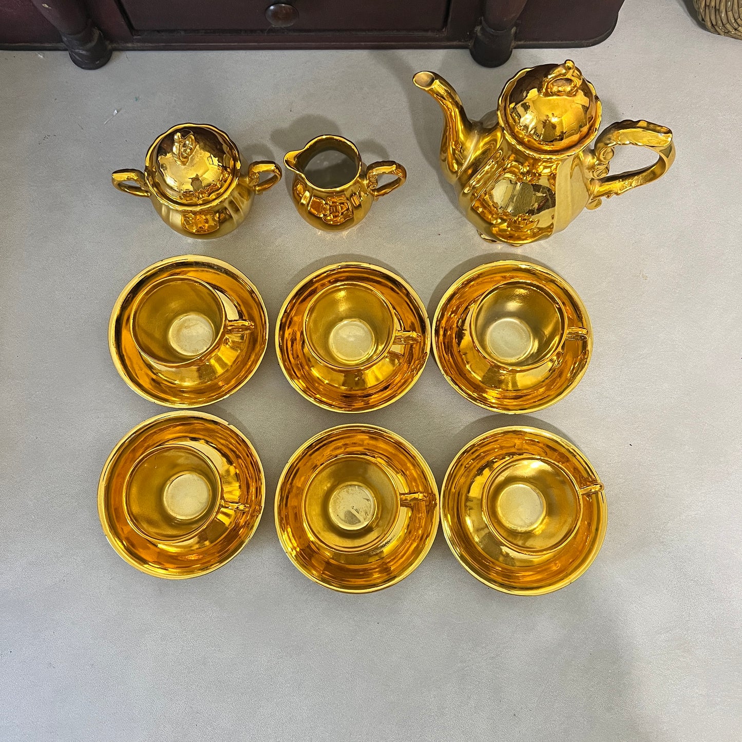 A gold-colored ceramic tea or coffee set arranged on a light gray floor, including a teapot, two small pitchers, and six matching cups with saucers.