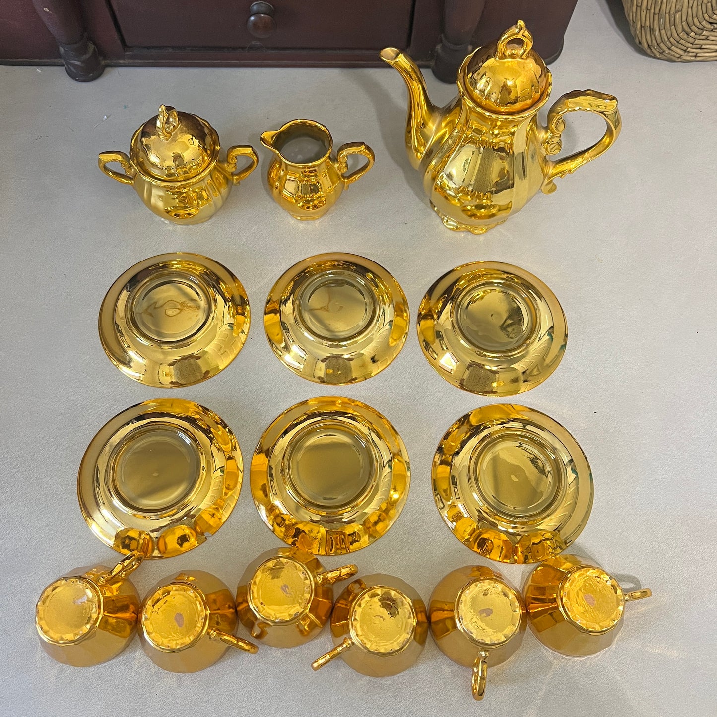 A set of shiny gold-colored ceramic tableware, including a teapot, teapot lid, creamer, four saucers, four cups, and four small bowls, arranged on a light gray surface.