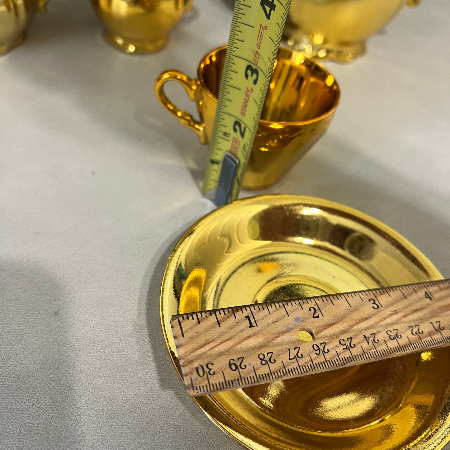 A shiny gold ceramic teacup and saucer set with a wooden ruler and a metal tape measure showing dimensions, photographed on a light gray surface.