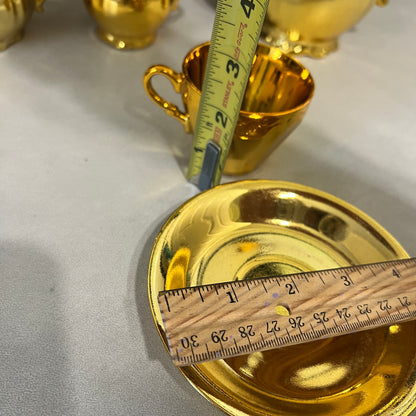 A shiny gold ceramic teacup and saucer set with a wooden ruler and a metal tape measure showing dimensions, photographed on a light gray surface.