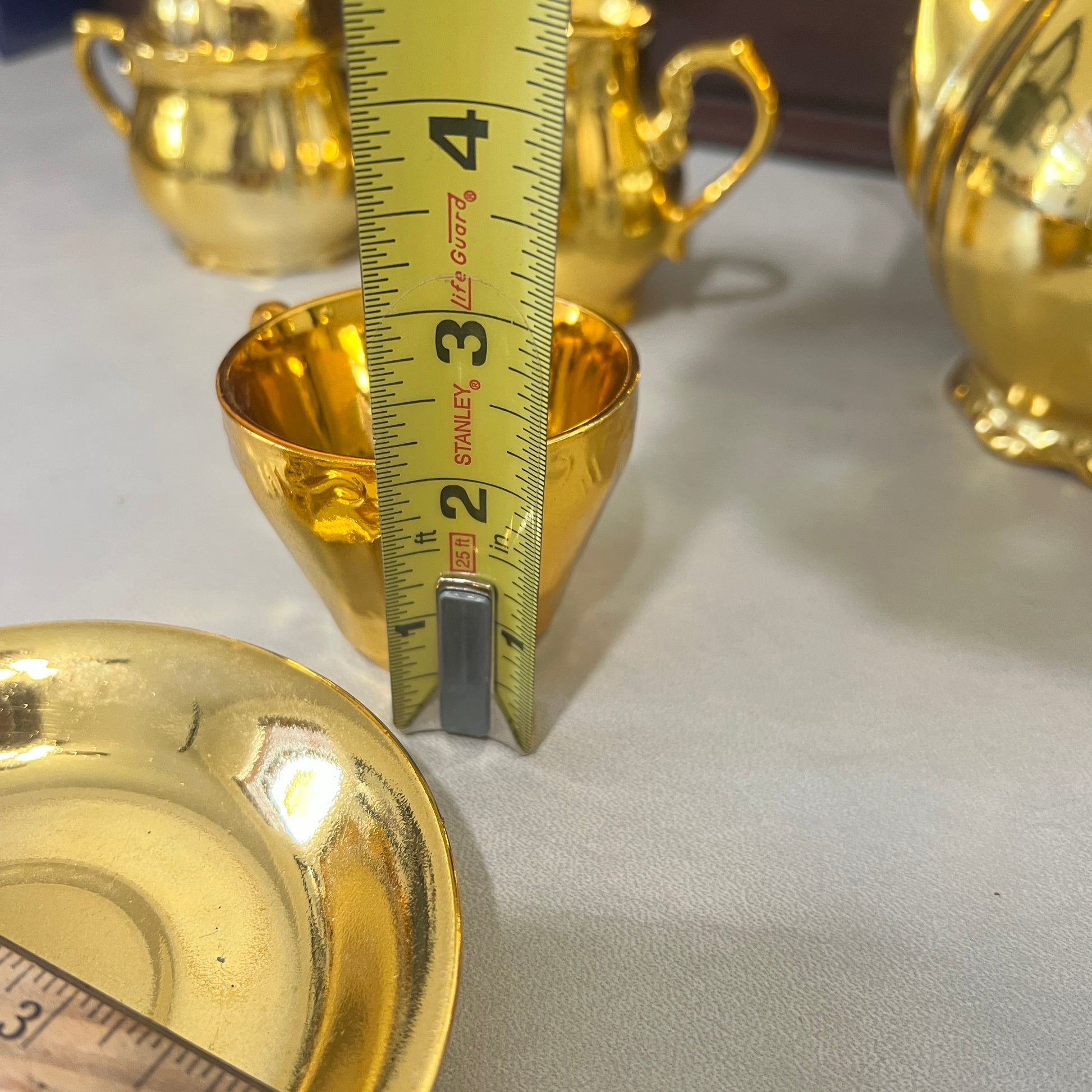 A close-up of a small gold cup or teacup with a yellow tape measure standing inside it to show the depth, on a light tabletop with other gold tableware in the background.