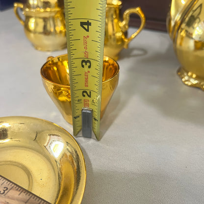 A close-up of a small gold cup or teacup with a yellow tape measure standing inside it to show the depth, on a light tabletop with other gold tableware in the background.