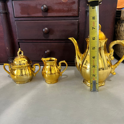 A shiny gold teapot with a yellow tape measure standing next to it, alongside two matching gold sugar bowls and a creamer, set against a dark wooden drawer background.