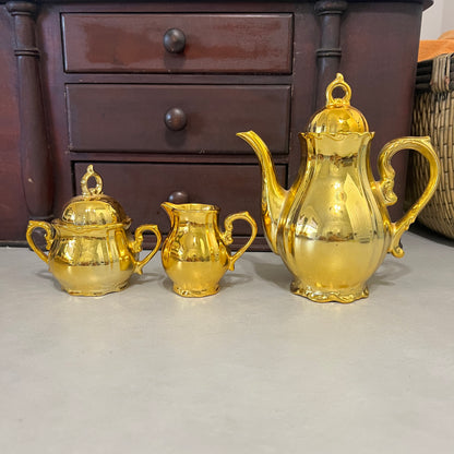 A set of three shiny gold ceramic tableware pieces, including a teapot, sugar bowl, and creamer, arranged on a light gray surface in front of a dark wooden chest of drawers.