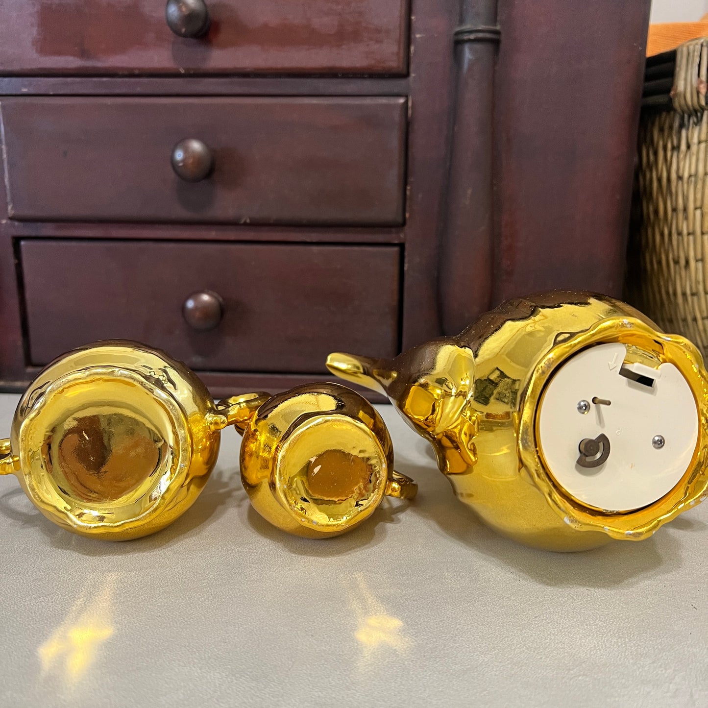 Three shiny gold ceramic tea or coffee cups arranged on a light surface in front of a dark wooden cabinet.