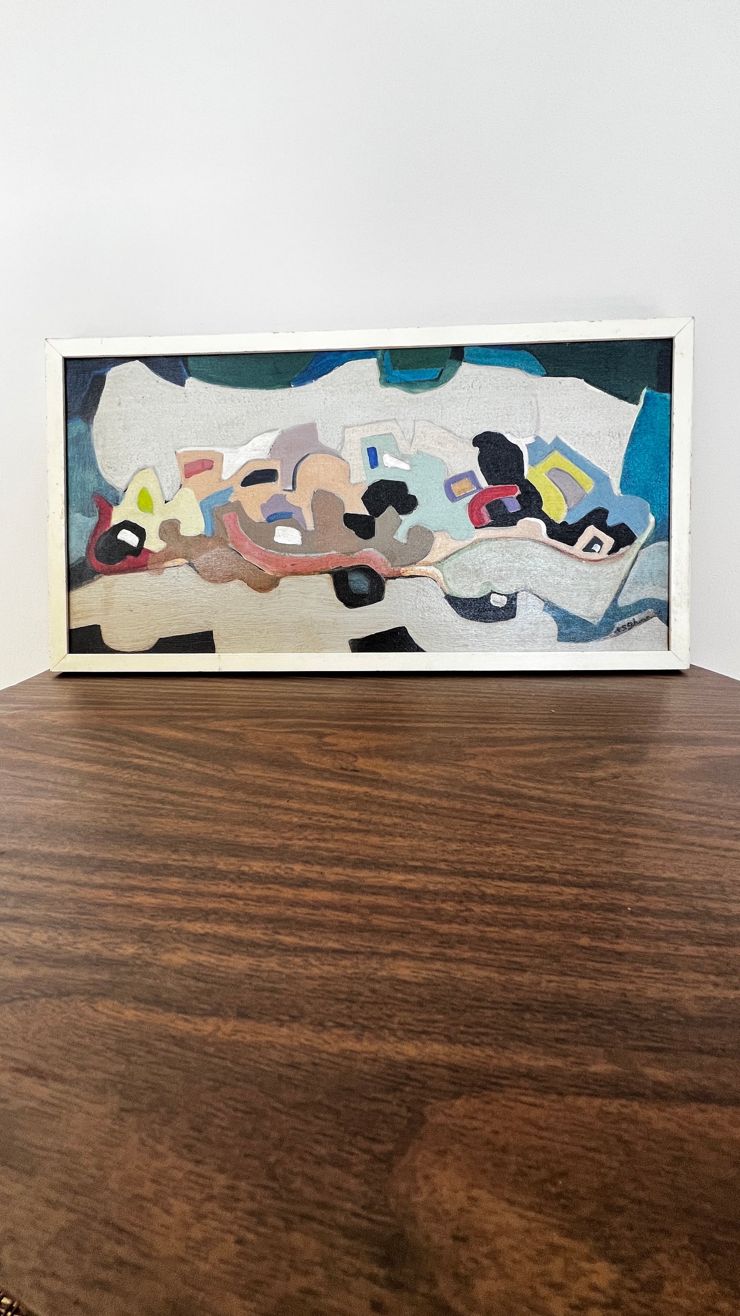 A framed abstract painting with a white frame, resting on a dark wood table against a plain white wall.