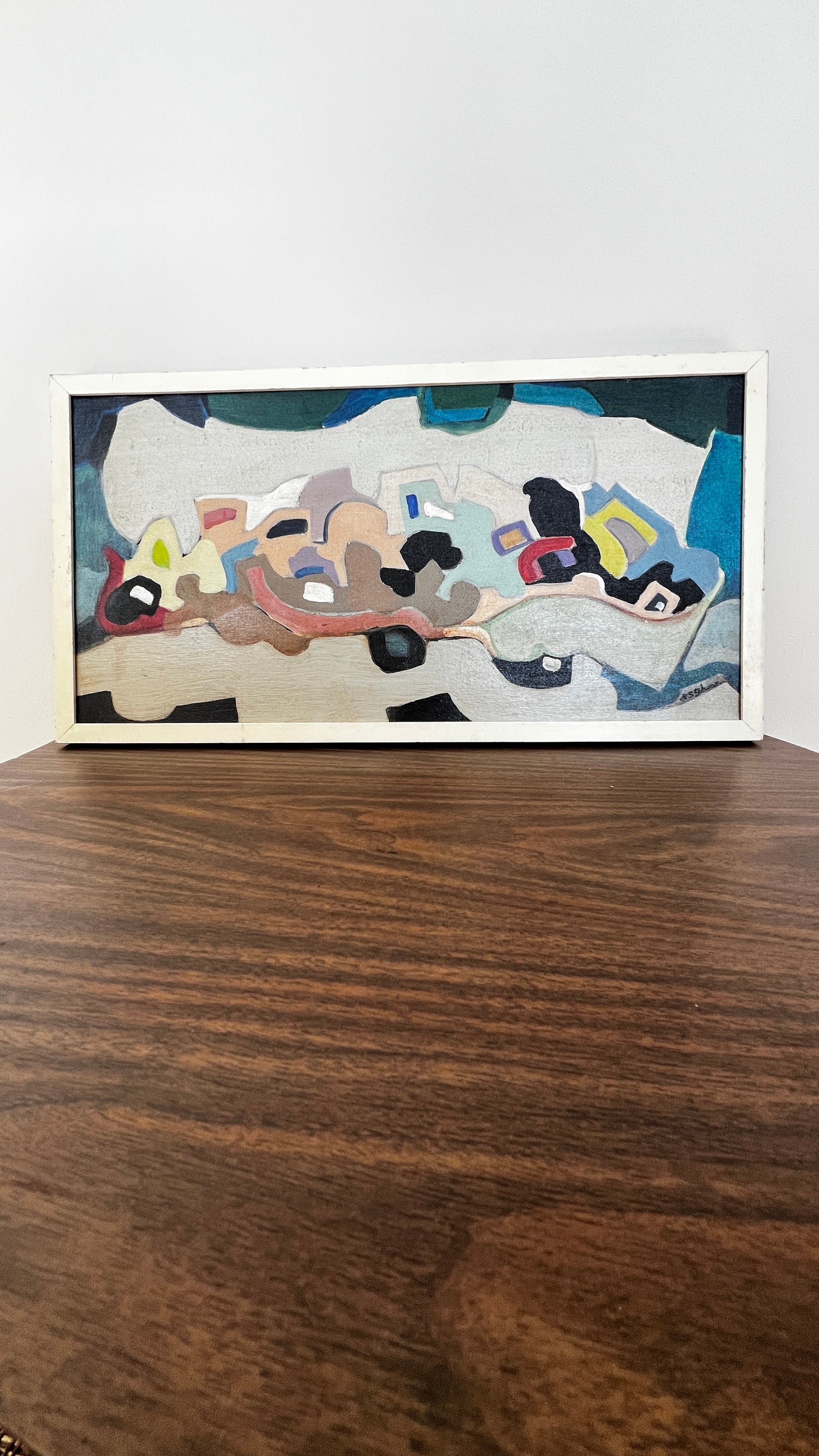 A framed abstract painting with a white frame, resting on a dark wood table against a plain white wall.