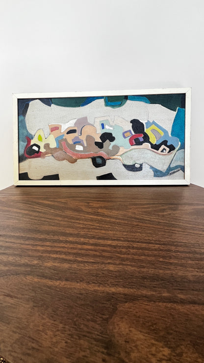 A framed abstract painting with a white frame, resting on a dark wood table against a plain white wall.