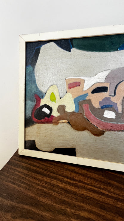 A framed abstract painting resting on a dark wood surface against a plain white wall. The artwork features layered, organic shapes in beige, brown, red, blue, green, and black, enclosed in a light-colored wooden frame.