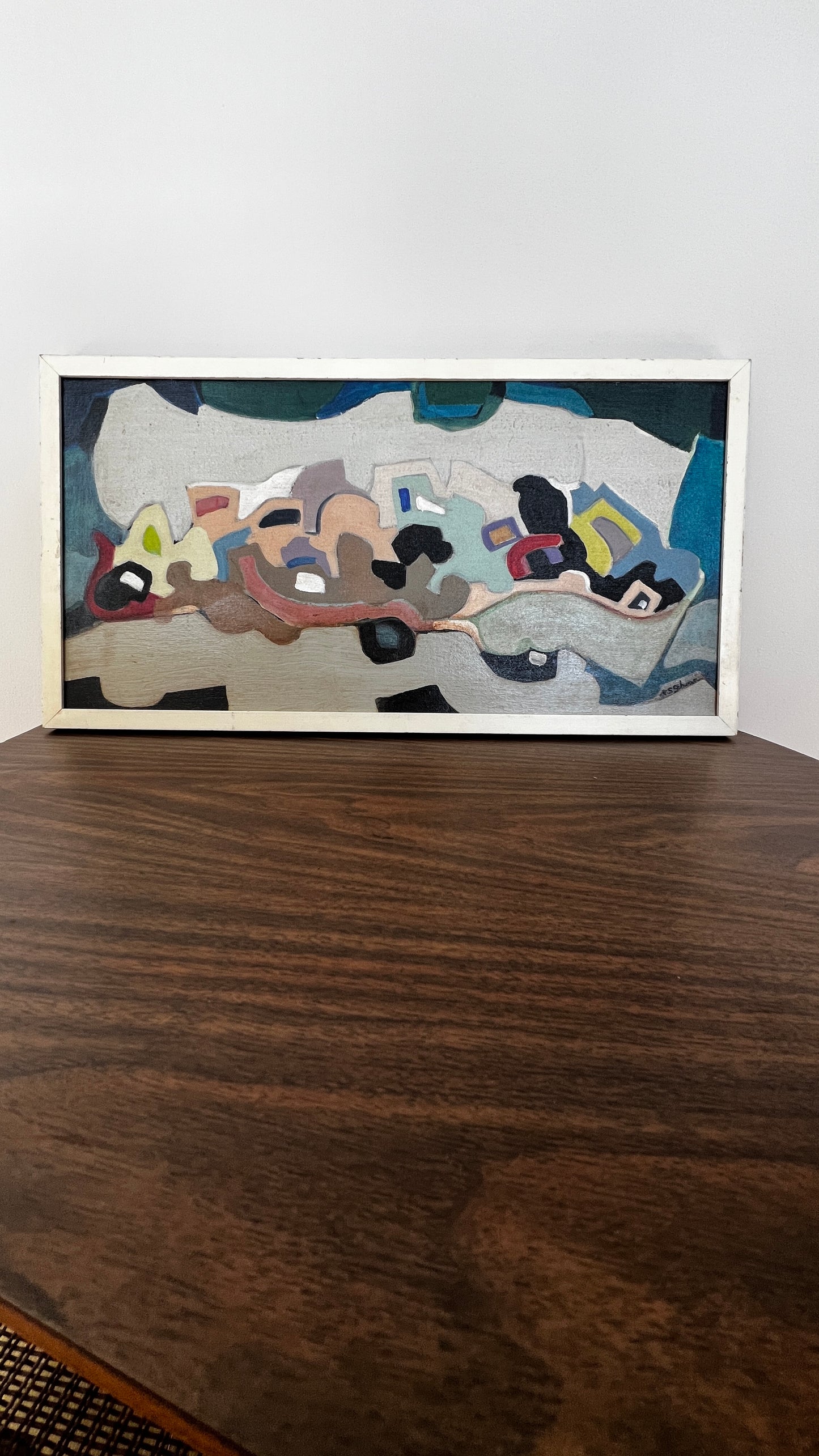 A framed abstract painting with a white frame, featuring a mix of gray, blue, black, brown, and beige shapes, displayed on a dark wooden table against a white wall.