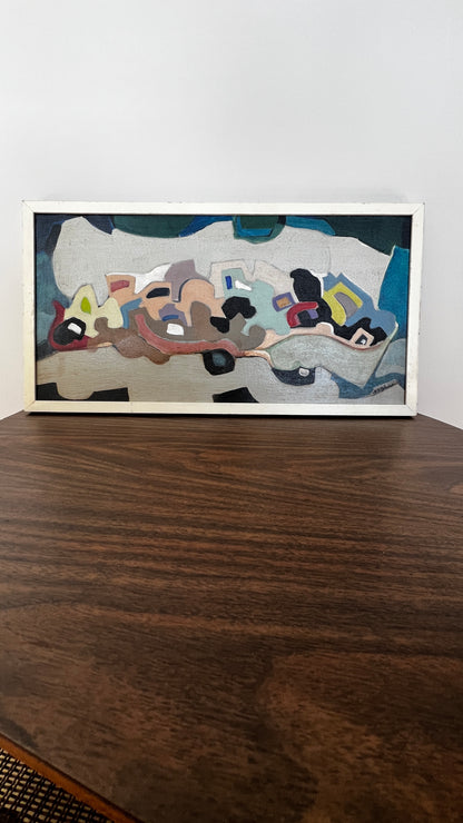 A framed abstract painting with a white frame, featuring a mix of gray, blue, black, brown, and beige shapes, displayed on a dark wooden table against a white wall.