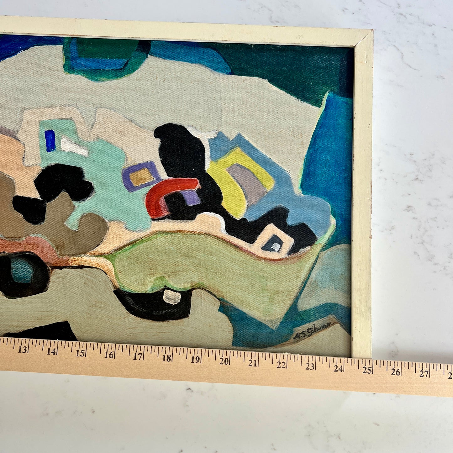 A framed abstract painting with geometric shapes in blue, green, beige, black, yellow, and red, shown on a white marble surface with a wooden ruler along the bottom edge for scale.