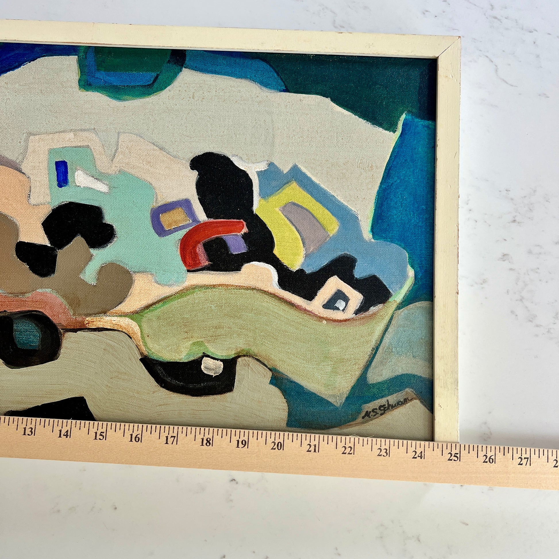 A framed abstract painting with geometric shapes in blue, green, beige, black, yellow, and red, shown on a white marble surface with a wooden ruler along the bottom edge for scale.