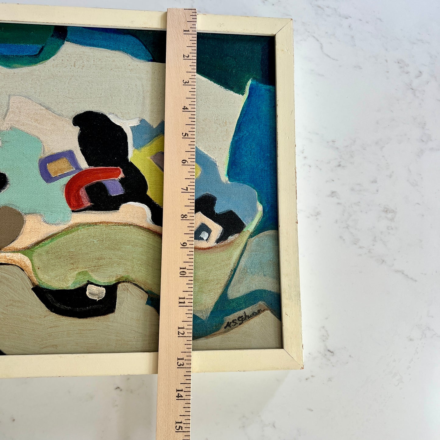 Abstract framed painting displayed on a white marble surface with a wooden ruler standing vertically to show scale. The painting features abstract shapes in teal, beige, black, red, and purple within a light wood frame.