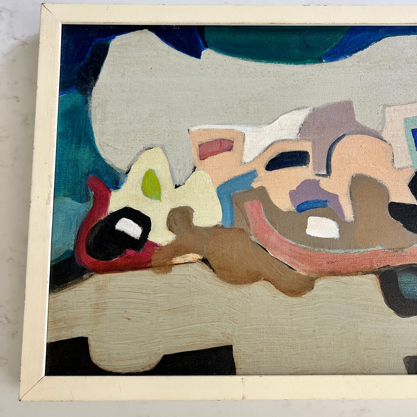A framed abstract painting featuring organic shapes in beige, brown, red, blue, green, and black, housed in a light cream-colored wooden frame.