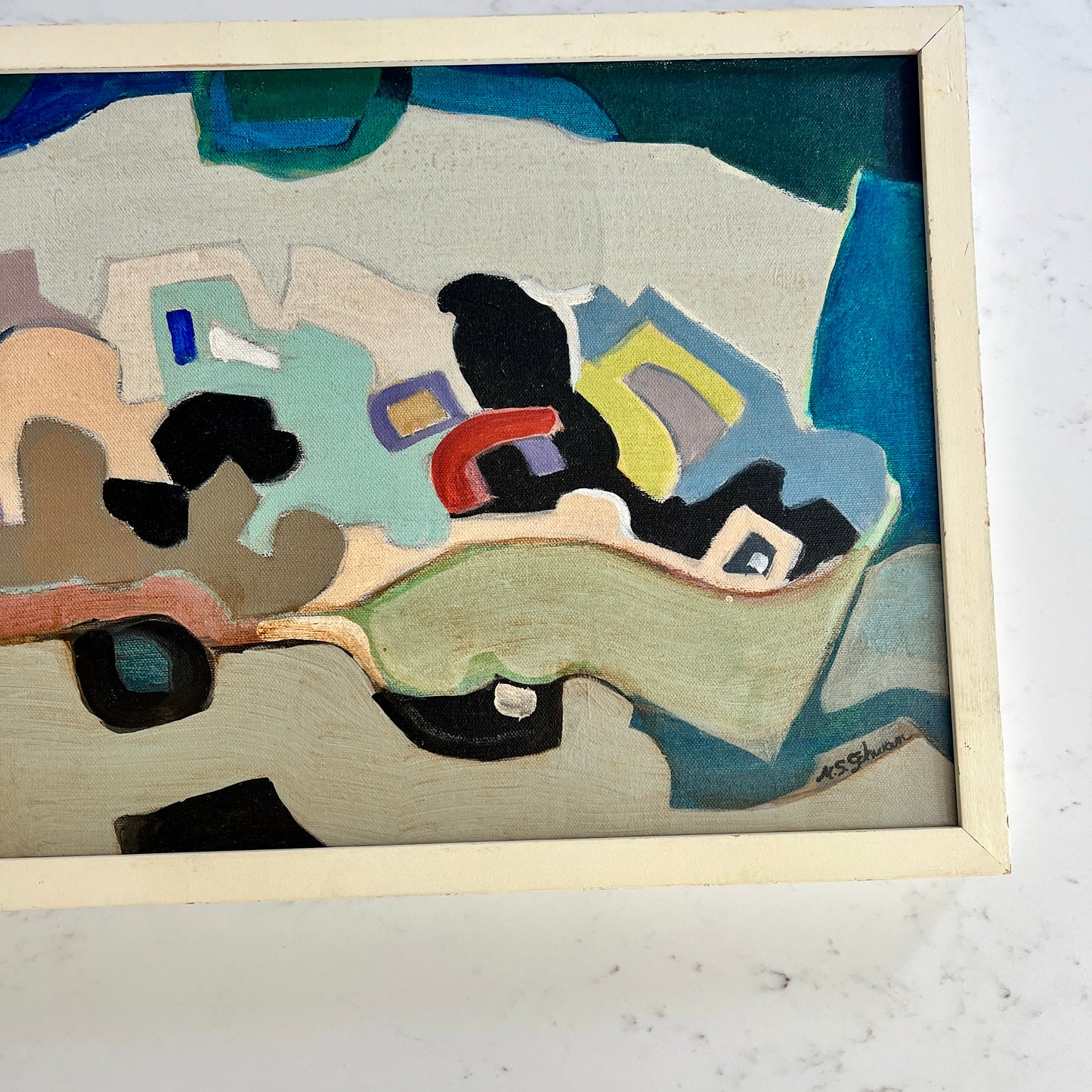 An abstract framed painting featuring organic shapes in shades of blue, beige, black, green, yellow, and red, housed in a light-colored wooden frame.