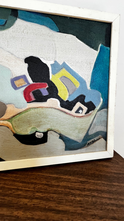 A framed abstract painting with bold geometric shapes in black, blue, white, yellow, red, and beige, resting on a dark wooden surface.