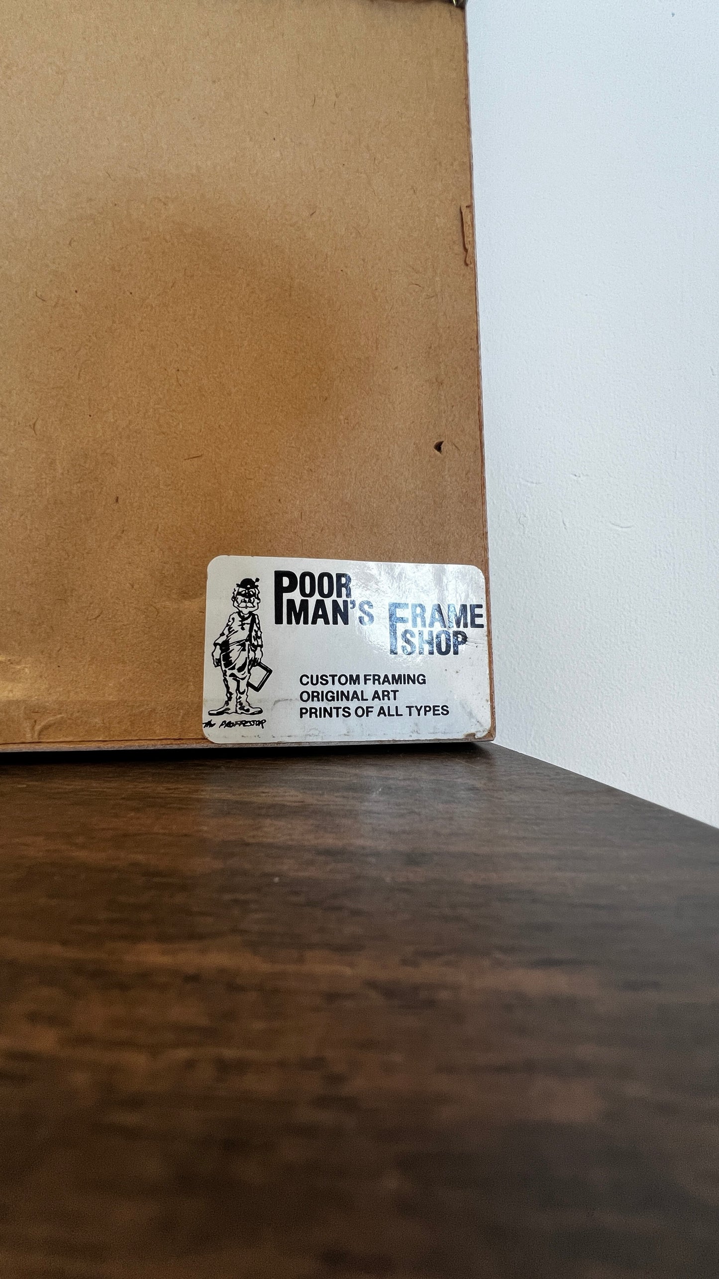 A white rectangular sticker on a brown cardboard box corner, reading 'POOR IMAN'S FRAME SHOP' with a small black line drawing of a person holding a frame.