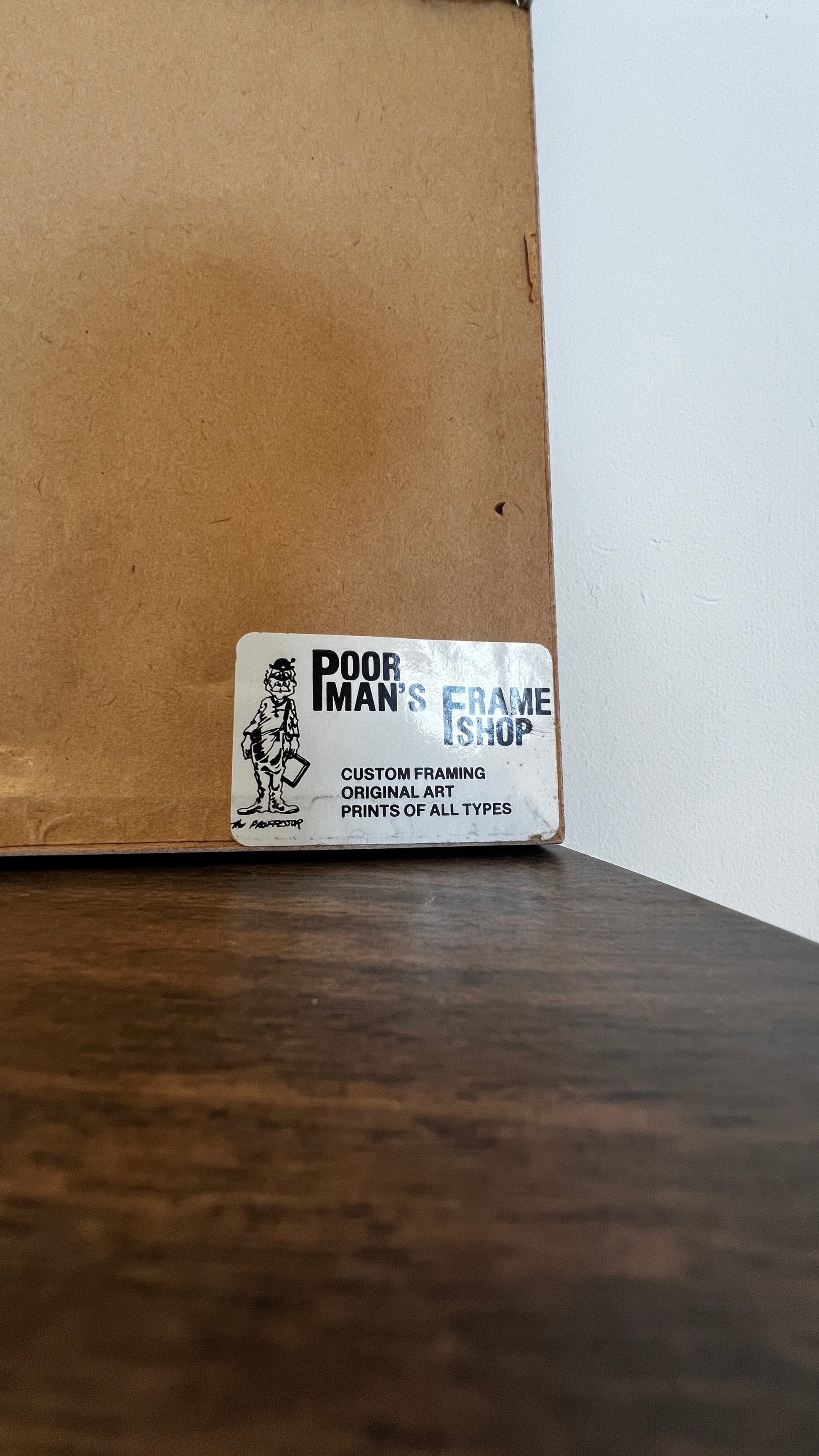 A white rectangular sticker on a brown cardboard box corner, reading 'POOR IMAN'S FRAME SHOP' with a small black line drawing of a person holding a frame.