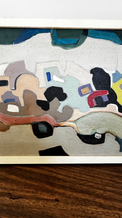 An abstract expressionist painting on textured canvas, featuring organic shapes and blocks of color including beige, black, blue, red, and gray, displayed on a dark wood surface.