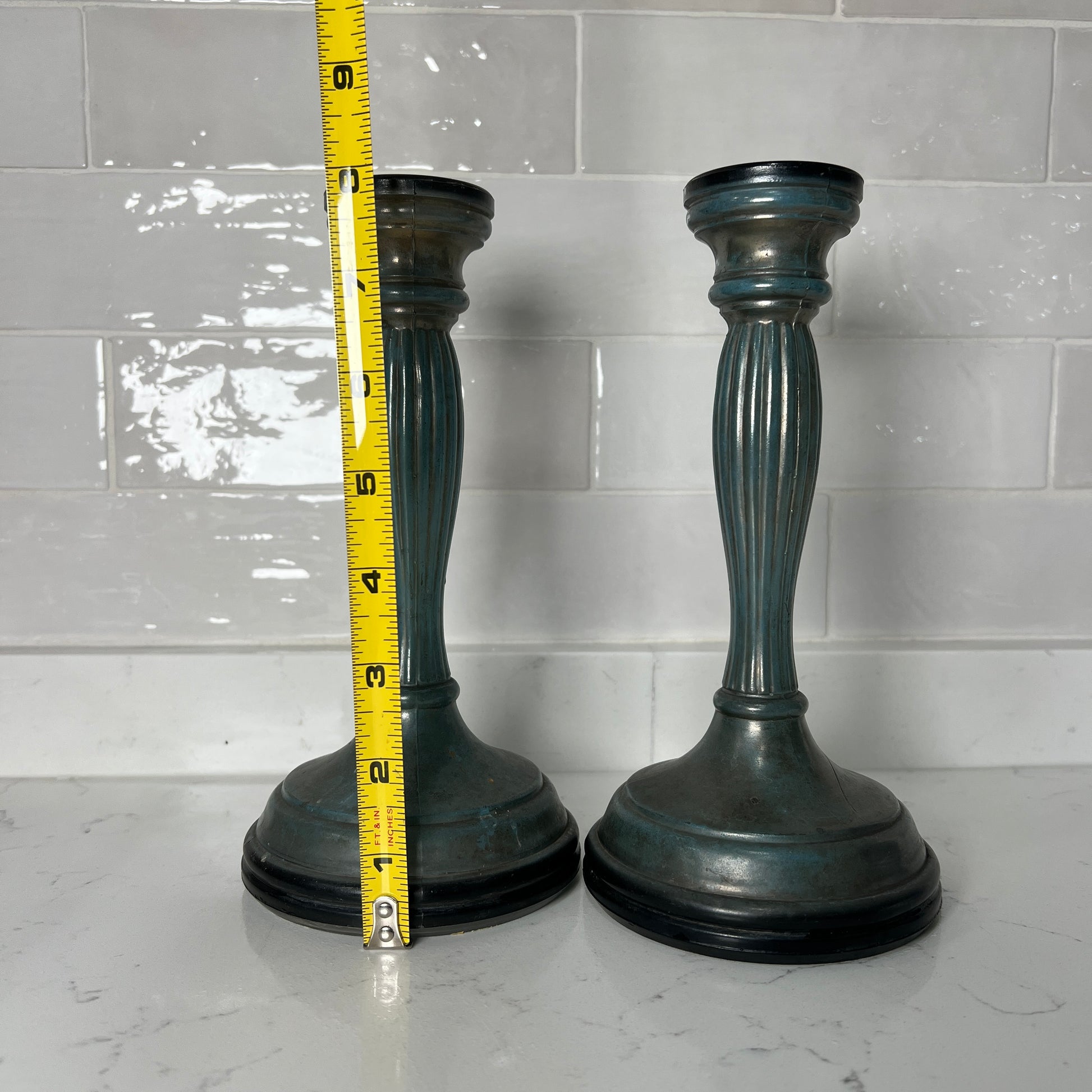 Two dark candle holders with a tape measure for scale against a tiled wall.