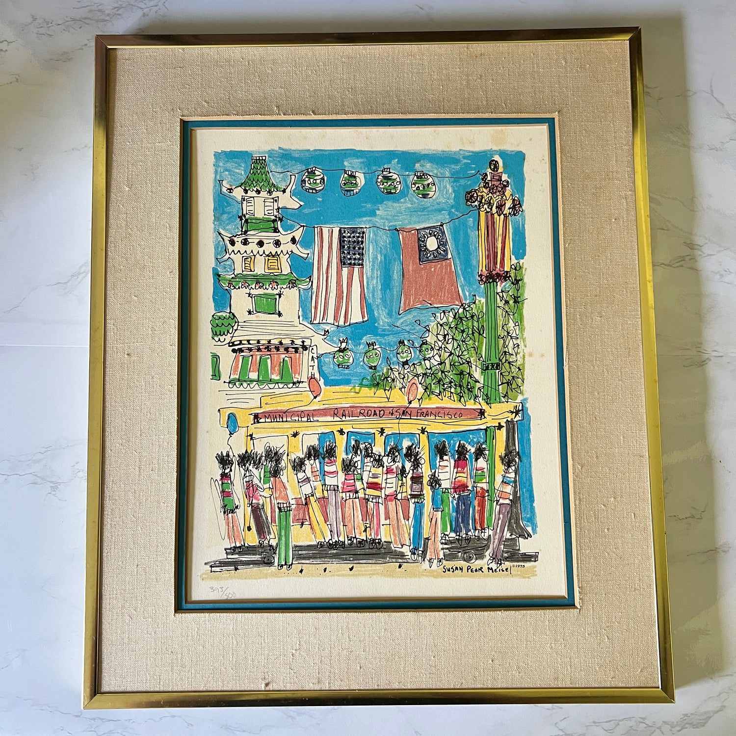 A framed folk art print depicting a street scene with a trolley, flags, and figures, mounted in a gold frame with a beige textured mat on a light marble surface.